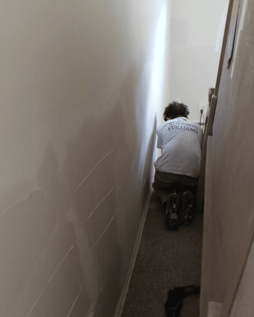 Worker wearing a Sherwin Williams shirt kneels in a narrow space patching a wall.