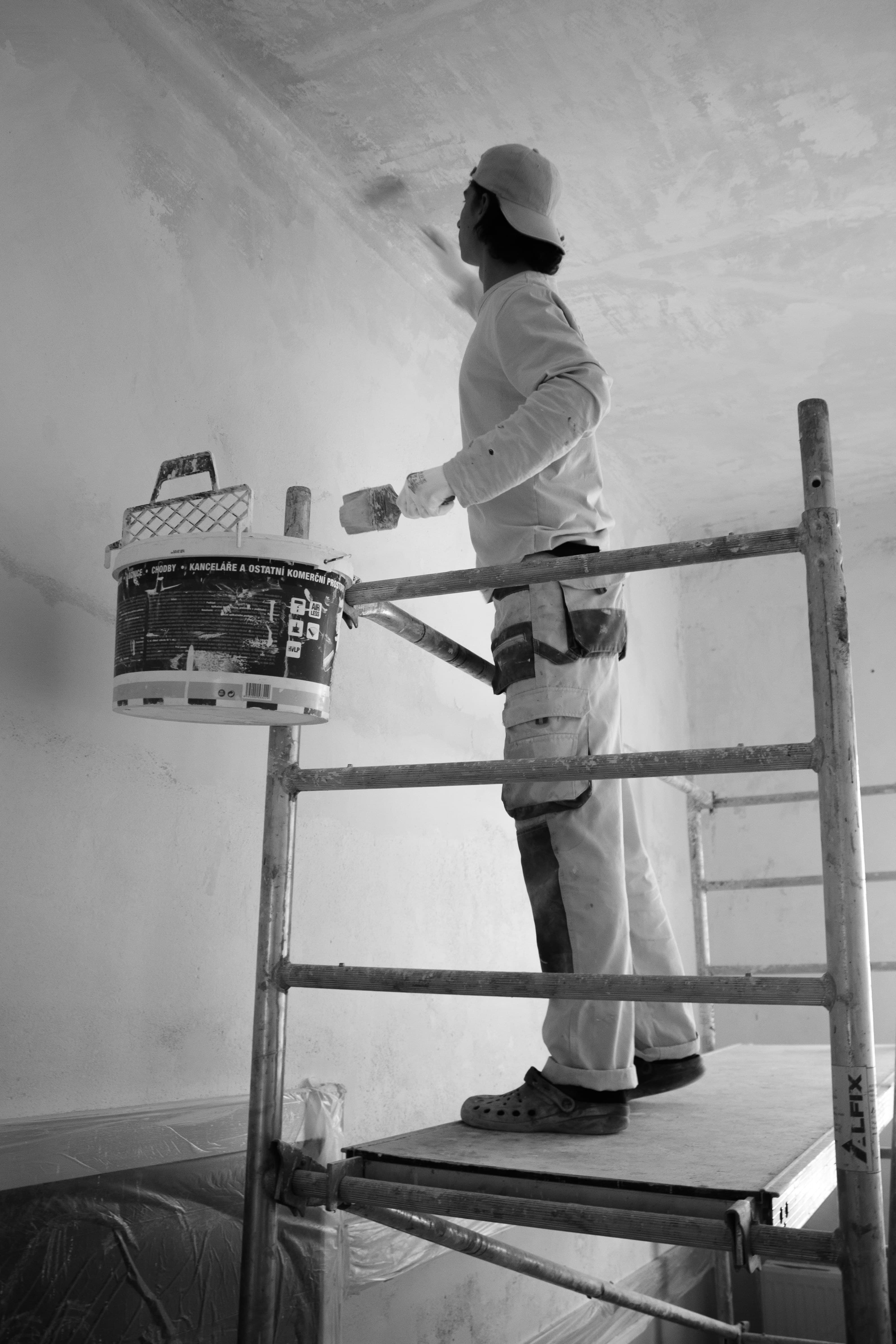 A worker on scaffolding paints a high wall with a brush next to a hanging bucket.