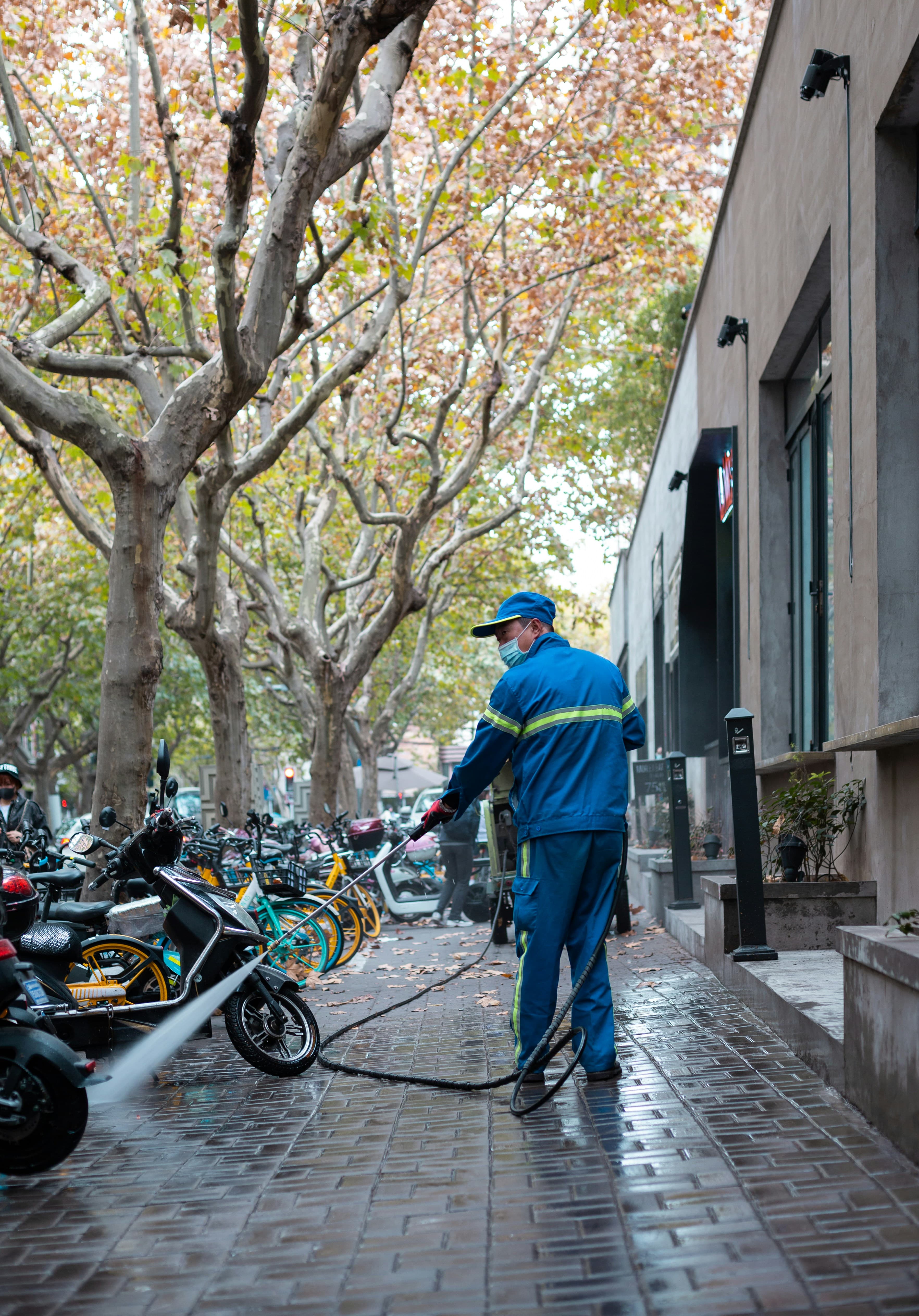 Sanitation worker in blue uniform power washes a sidewalk lined with bicycles and autumn trees.